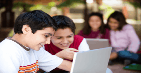 Two primary-school boys smiling as they share a laptop outdoors, with two girls sitting behind them